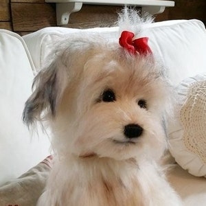 May include: A white fluffy dog with a red bow on its head sits on a white sofa. The dog has a white and grey coat and is looking at the camera.