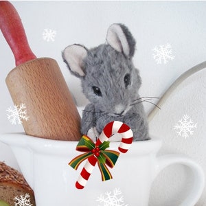 Puede incluir: Un ratón gris con un bastón de caramelo en sus patas está sentado en una taza blanca con un lazo rojo y verde. La taza está decorada con copos de nieve. El texto "Merry Christmas" y "Teddy-Manufaktur.de" está impreso en la parte inferior de la imagen.