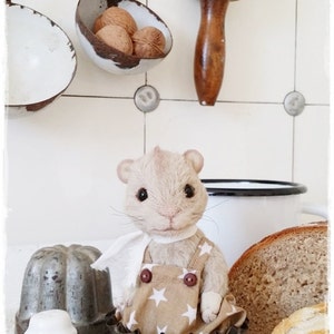 May include: A small, plush, tan-colored mouse wearing overalls with white stars and a white scarf sits in a metal baking cup. The mouse is looking directly at the camera. The mouse is surrounded by other baking cups and a loaf of bread.