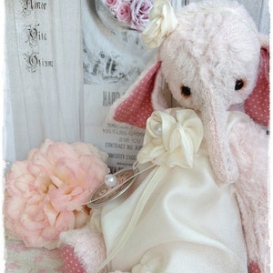 May include: A pink plush elephant toy wearing a white satin dress with a pink polka dot bow and a white satin ribbon. The elephant has a long trunk and large ears. The toy is sitting on a white surface with a floral pattern.