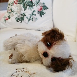May include: A white and brown plush toy dog with brown eyes and a white muzzle, lying on a white fluffy blanket with a floral pattern.
