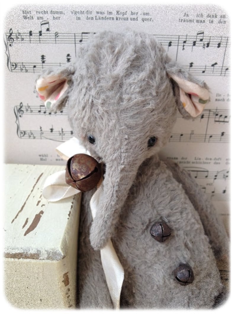 Puede incluir: Un juguete de elefante de peluche gris con una trompa larga y orejas grandes. El juguete tiene una cinta blanca atada alrededor de su cuello y tres peque&ntilde;as campanas oxidadas unidas a la cinta. El juguete est&aacute; sentado sobre una superficie de madera blanca.