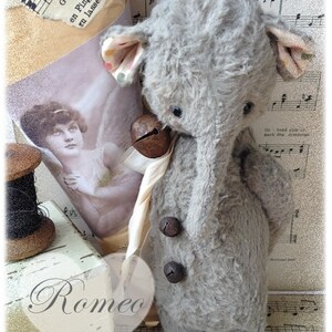 Puede incluir: Un juguete de elefante de peluche gris con una campana alrededor del cuello y dos botones en el pecho. El juguete est&aacute; sentado en una hoja de m&uacute;sica con la palabra "Romeo" escrita en un bloque de madera en primer plano.