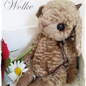 Könnte beinhalten: Ein brauner Teddybär mit großen schwarzen Augen und kleinen braunen Hörnern. Der Bär hat ein Band um den Hals und sitzt neben einer Gänseblümchen und einer roten Rose. Der Text "Schäfchen Wolke" steht über dem Bären.