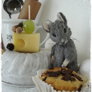 May include: A gray plush mouse figurine sitting on a white cake stand with a slice of cheese and a cupcake with chocolate chips on top.
