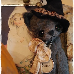 May include: A gray plush toy cat wearing a black witch hat with a brown and white patterned band. The cat has a white ribbon tied around its neck and a tag that says "Booh".
