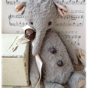 Puede incluir: Un juguete de elefante de peluche gris con una cinta blanca y dos campanas de metal. El juguete est&aacute; sentado en una caja de madera blanca con una hoja de m&uacute;sica en el fondo.