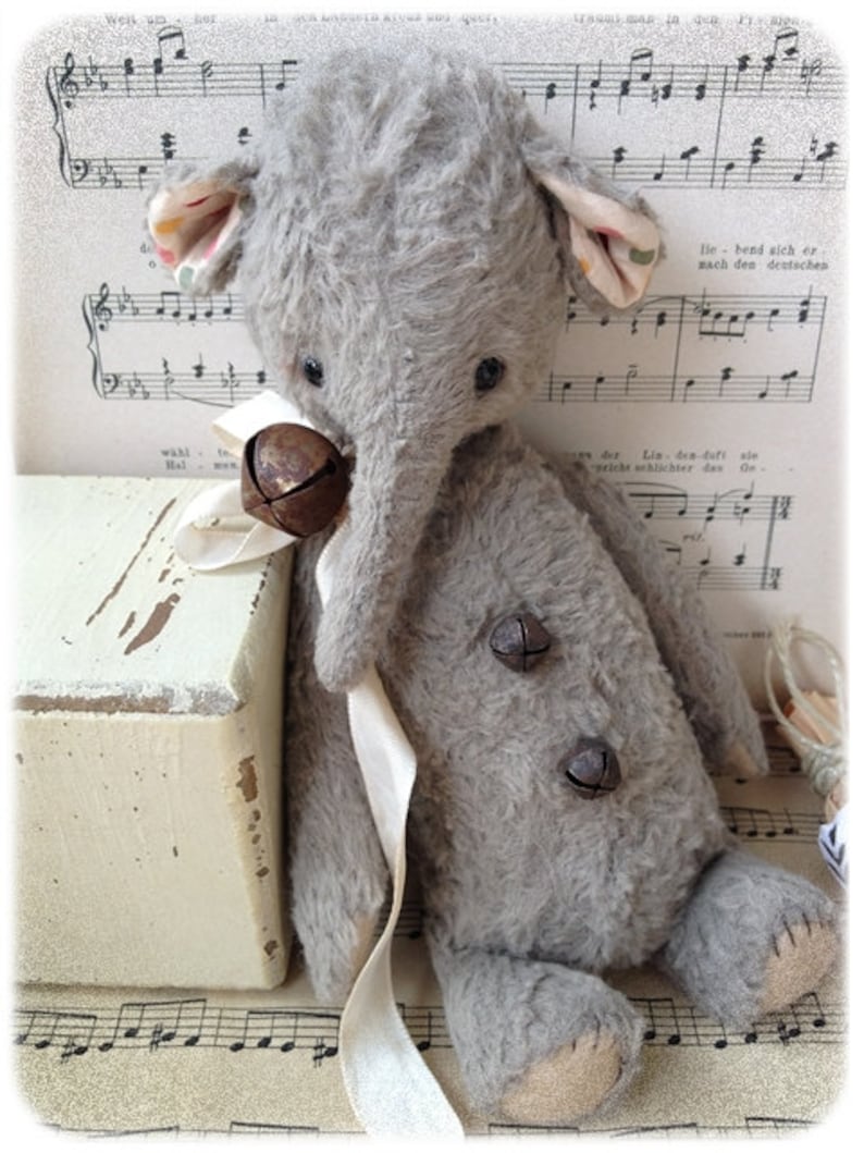 Puede incluir: Un juguete de elefante de peluche gris con una cinta blanca y dos campanas de metal. El juguete est&aacute; sentado en una caja de madera blanca con una hoja de m&uacute;sica en el fondo.