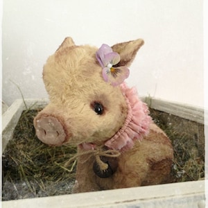 Peut inclure: Un jouet en peluche de cochon fait main avec un col à volants rose et une fleur violette sur la tête. Le cochon est assis dans une boîte en bois remplie de foin.