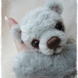 May include: A soft, gray plush teddy bear with brown button eyes and a nose. The bear is being held by a hand.