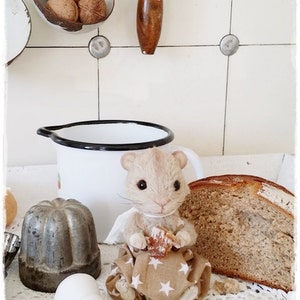 May include: A small, plush, light brown toy mouse wearing a brown and white star patterned dress, holding a small brown pastry. The mouse is sitting on a white crocheted surface with a brown loaf of bread in the background.