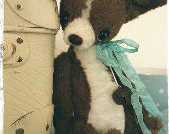 May include: A brown and white plush toy dog with a blue ribbon around its neck. The dog is sitting next to a vintage suitcase.