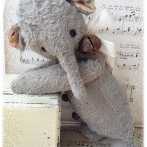 Puede incluir: Un juguete de elefante de peluche gris con una campana atada al cuello. El elefante est&aacute; sentado en una caja de madera con una superficie pintada de blanco. El juguete est&aacute; encima de una partitura musical con notas musicales.
