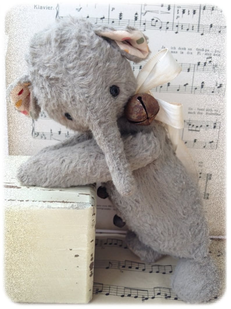 Puede incluir: Un juguete de elefante de peluche gris con una campana atada al cuello. El elefante est&aacute; sentado en una caja de madera con una superficie pintada de blanco. El juguete est&aacute; encima de una partitura musical con notas musicales.