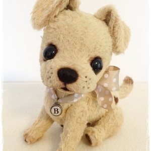 May include: A beige plush toy dog with brown eyes and a black nose. The dog is wearing a white collar with a brown polka dot ribbon and a silver tag with the letter 'B' on it.