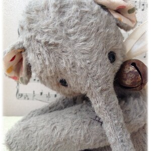 Puede incluir: Un juguete de elefante de peluche gris con una campana marr&oacute;n unida a su oreja. El elefante est&aacute; sentado sobre una superficie blanca con notas musicales en el fondo.