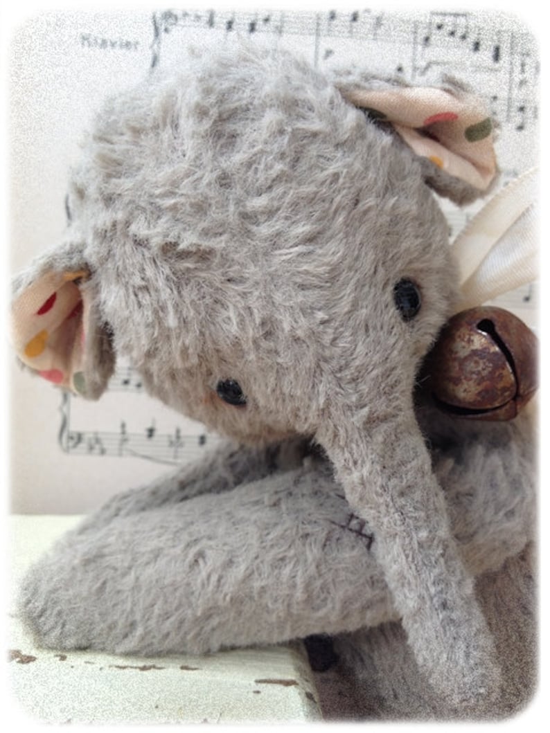 Puede incluir: Un juguete de elefante de peluche gris con una campana marr&oacute;n unida a su oreja. El elefante est&aacute; sentado sobre una superficie blanca con notas musicales en el fondo.