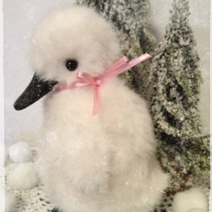 Pode incluir: Um brinquedo de pelúcia de pato branco e felpudo com uma fita rosa em volta do pescoço. O pato está sentado em uma superfície de crochê branca com bolas de lã brancas no fundo.