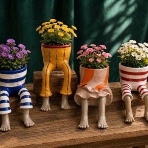 Pode incluir: Quatro vasos de cerâmica em forma de parte inferior do corpo com pernas e pés, cada um contendo uma planta com flores diferente. Os vasos apresentam vários designs, incluindo listras azuis e brancas, amarelo sólido, laranja e bege.