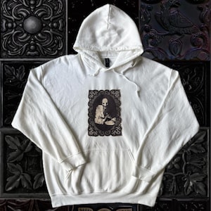 May include: A white hooded sweatshirt featuring a central rectangular graphic print of a figure in a dark, ornate frame. The garment includes long sleeves, a front kangaroo pocket, and matching white drawstring cords attached to the hood.