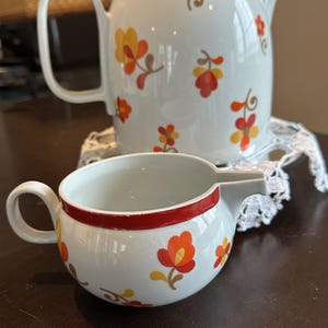 May include: A white ceramic teapot and matching creamer featuring a repeating floral pattern in shades of orange, yellow, and red. The teapot has a lid with a red rim, and the creamer features a red band around the top rim and a small spout.