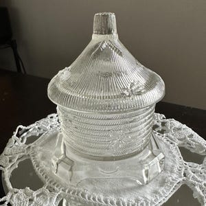 May include: A clear glass honey pot shaped like a beehive with a matching lid. The glass features a textured, ribbed surface pattern resembling woven straw and is decorated with embossed bee figures on both the lid and the main body section.
