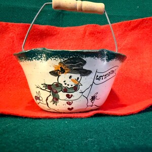 May include: A white ceramic bowl with a dark green rim and a metal handle featuring a wooden grip. The bowl is decorated with a hand-painted snowman wearing a black hat and a green scarf, holding a flag with the text LET IT SNOW written on it.