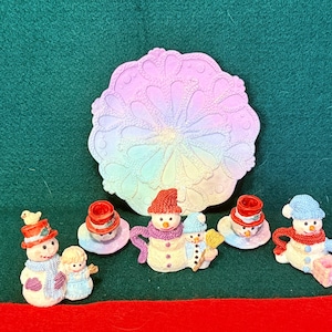 May include: A collection of miniature snowman figurines and a circular decorative plate. The plate features a pastel gradient of pink, blue, and yellow with an embossed floral pattern. The snowmen wear colorful hats, scarves, and mittens.