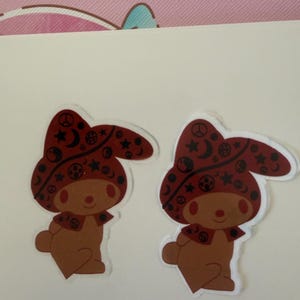 May include: Two identical stickers featuring a brown cartoon rabbit character wearing a dark red hood. The hood is decorated with black symbols including stars, crescent moons, peace signs, and yin-yang icons. The rabbit has a simple, rounded face.
