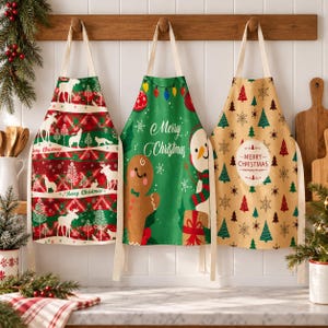 May include: Three kitchen aprons hanging on a wooden rack. The left apron features a red and green plaid pattern with white moose and trees. The center green apron displays a gingerbread man, a snowman, and the text Merry Christmas. The right tan apron has a repeating tree pattern and a circular Merry Christmas label.