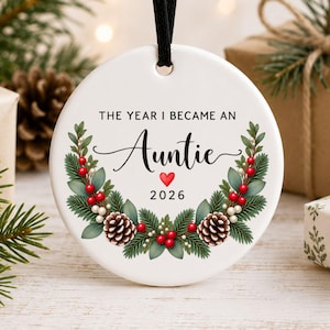 Op de afbeelding: Een rond wit keramieken ornament hangend aan een zwart lint. De tekst luidt THE YEAR I BECAME AN Auntie 2026 met een klein rood hartje. Een krans van groene dennentakken, bruine dennenappels en rode bessen omlijst de tekst op het ornament.