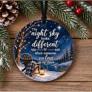 May include: A round ceramic ornament featuring a night sky scene with a wooden chair, a plaid blanket, and a lantern on a snowy shore. The text reads: The night sky looks different when someone you love is up there. It hangs from a black ribbon.