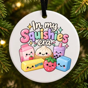 Könnte beinhalten: Ein rundes weißes Keramikornament mit dem Text In my Squishies era in bunten Buchstaben. Unter dem Text befinden sich niedliche Cartoon-Figuren, darunter ein rosa Milchkarton, ein Teigtäschchen, ein Bubble Tea, ein Stück Butter, eine Erdbeere und ein blauer Würfel.