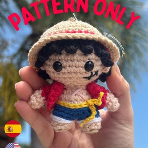 Może przedstawiać: Mała szydełkowa lalka amigurumi z beżowym słomkowym kapeluszem z czerwoną opaską, czarnymi włosami i wyszywaną blizną pod lewym okiem. Lalka ma na sobie czerwoną rozpiętą kamizelkę, niebieskie szorty z żółtym pasem oraz widoczny napis PATTERN ONLY.