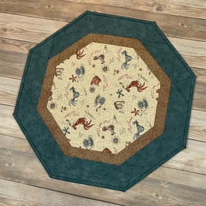 May include: An octagonal fabric mat featuring a central cream-colored panel with a repeating pattern of roosters, flowers, and the text "Rise and Shine." The mat has a brown inner border and a wide, dark teal outer border with subtle stitching.
