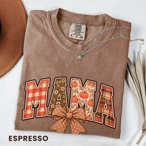 Puede incluir: Una camiseta marrón de manga corta con cuello redondo y etiqueta Comfort Colors. El frente presenta el texto MAMA en letras grandes con patrones de cuadros, flores y calabazas, acentuado por un gráfico de lazo marrón con lunares en la base.