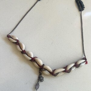 May include: A seashell hair accessory featuring cream-colored shells strung together with dark red cord and a dark metal chain. The accessory has two black hair clips on each end and a dangling charm.