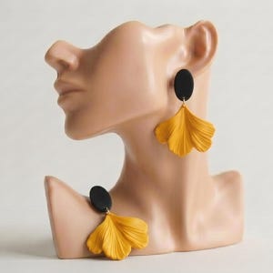 May include: A pair of dangle earrings featuring a matte black oval stud attached to a textured, fan-shaped mustard yellow pendant. The earrings are displayed on a beige mannequin bust, showcasing the contrast between the dark oval top and the bright, pleated leaf-like design of the lower portion of the jewelry piece.