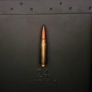 May include: A single rifle cartridge with a copper-coloured pointed bullet and a brass casing, positioned vertically against a dark olive green metal ammunition box that features the embossed text 24 M2A1 on its front surface.