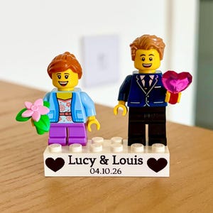 May include: Two plastic minifigures standing on a white rectangular base printed with the text Lucy & Louis and the date 04.10.26, flanked by two black heart icons. The left figure has brown hair, a blue jacket, and purple trousers, holding a pink flower. The right figure has brown hair, a dark blue suit, and black trousers, holding a translucent pink heart.