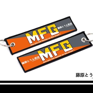 May include: Two rectangular keychains featuring a black border, orange and grey geometric background, and the bold white text MFG. The text reads Are you the fastest on the street? and includes the Japanese characters for Fujiwara Tofu Cafe.