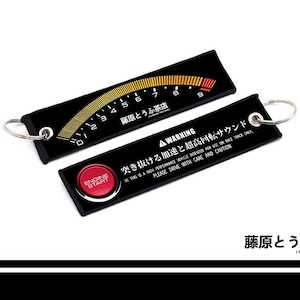 May include: Two rectangular black fabric keychains with metal rings. One features a tachometer gauge graphic with numbers zero through nine and Japanese text. The other displays a red engine start button, a warning label, and Japanese text.