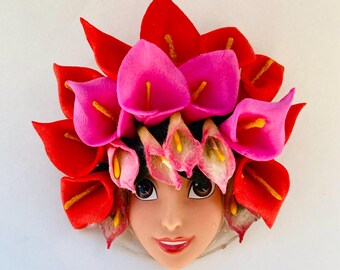 Puede incluir: Cabeza de muñeca adornada con un tocado floral. El tocado presenta calas rojas y rosas vibrantes con estambres amarillos. La muñeca tiene ojos oscuros y una expresión sonriente. El diseño es colorido y decorativo.