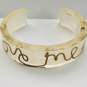 Könnte beinhalten: Transparenter, offener Armreif mit den Worten "love me" in goldfarbener Schrift. Das Armband besteht aus einem durchscheinenden Material mit leicht gealtertem Aussehen. Ein einzigartiges Schmuckstück.