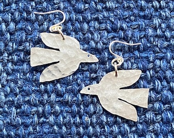 Könnte beinhalten: Ein Paar silberfarbene Ohrringe in Vogelform mit gehämmerten Texturen. Die Ohrringe haben einen Hakenverschluss und werden auf einer blauen, strukturierten Oberfläche präsentiert. Die Vögel haben einen kleinen blauen Punkt für das Auge.