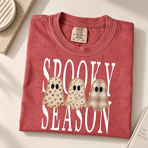 May include: A brick red t-shirt featuring the white text SPOOKY SEASON and three ghost illustrations. The ghosts have distinct patterns: one with brown hearts, one with white daisies, and one with a tan plaid design, all with black oval eyes.