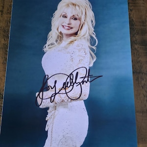 May include: A glossy photograph featuring a portrait of Dolly Parton with blonde hair, wearing a white lace long-sleeved dress against a blue background. The image includes a prominent black ink autograph signature across the centre of the print.