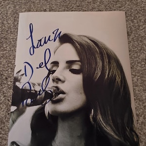 Puede incluir: Una impresión fotográfica en blanco y negro que muestra a una persona bebiendo de una botella de vidrio, superpuesta con una firma en tinta azul que dice Lana Del Rey. El retrato captura al sujeto desde el pecho hacia arriba de perfil.