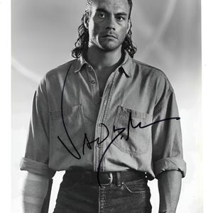 Peut inclure: Une photographie promotionnelle en noir et blanc montrant une personne avec une coupe mulet, portant une chemise boutonnée aux manches retroussées et une ceinture. L'image comporte une signature à l'encre noire et un texte de copyright en bas.