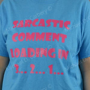 May include: A bright blue short-sleeved t-shirt featuring bold pink text that reads SARCASTIC COMMENT LOADING IN 3... 2... 1... The lettering is centered on the chest area of the garment, creating a clear and readable graphic design on the fabric.